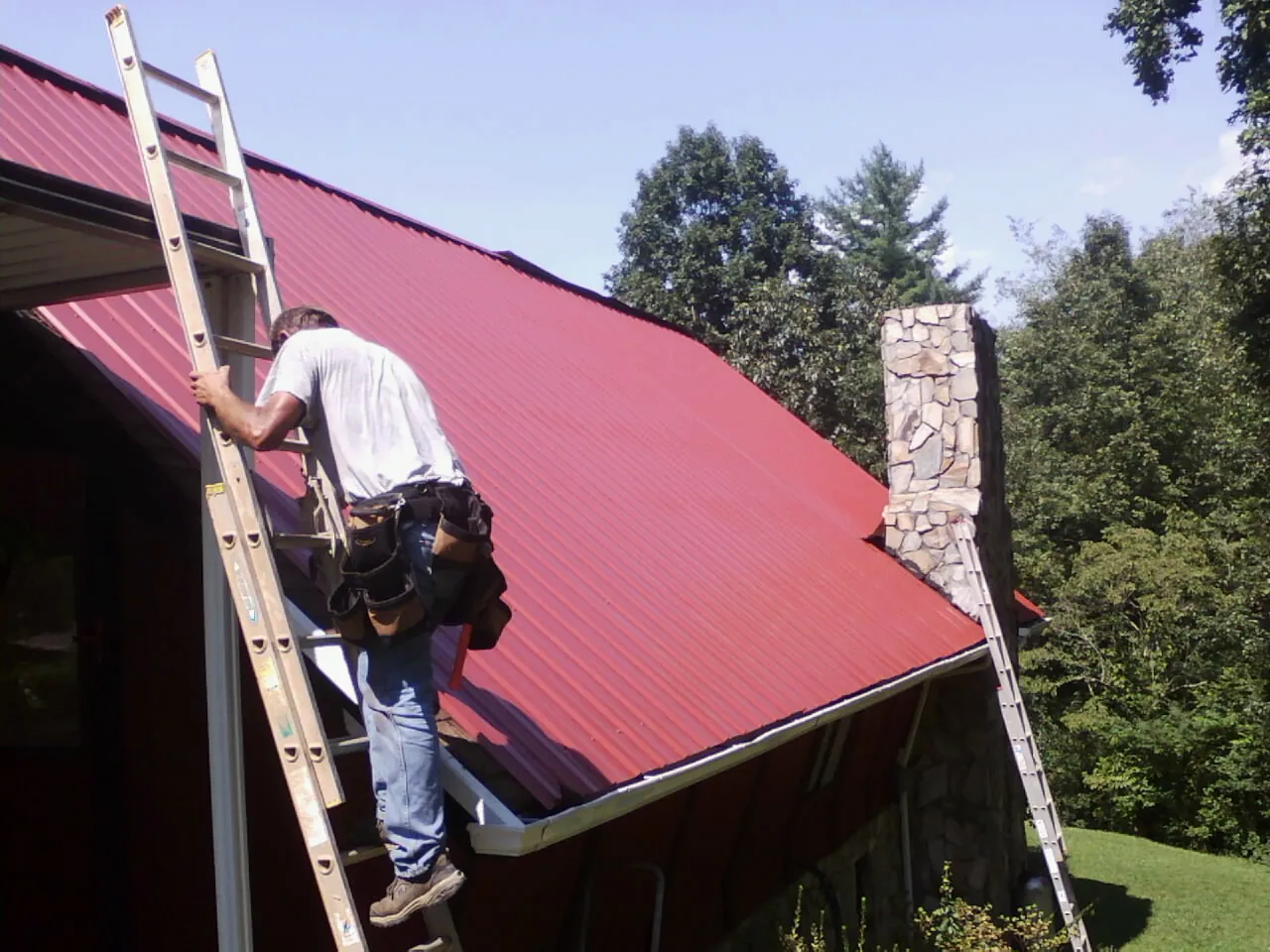 Roofer on a ladder beside a red metal roof and stone chimney — Soffit & Fascia Repair by Roofing Brothers in Dobson